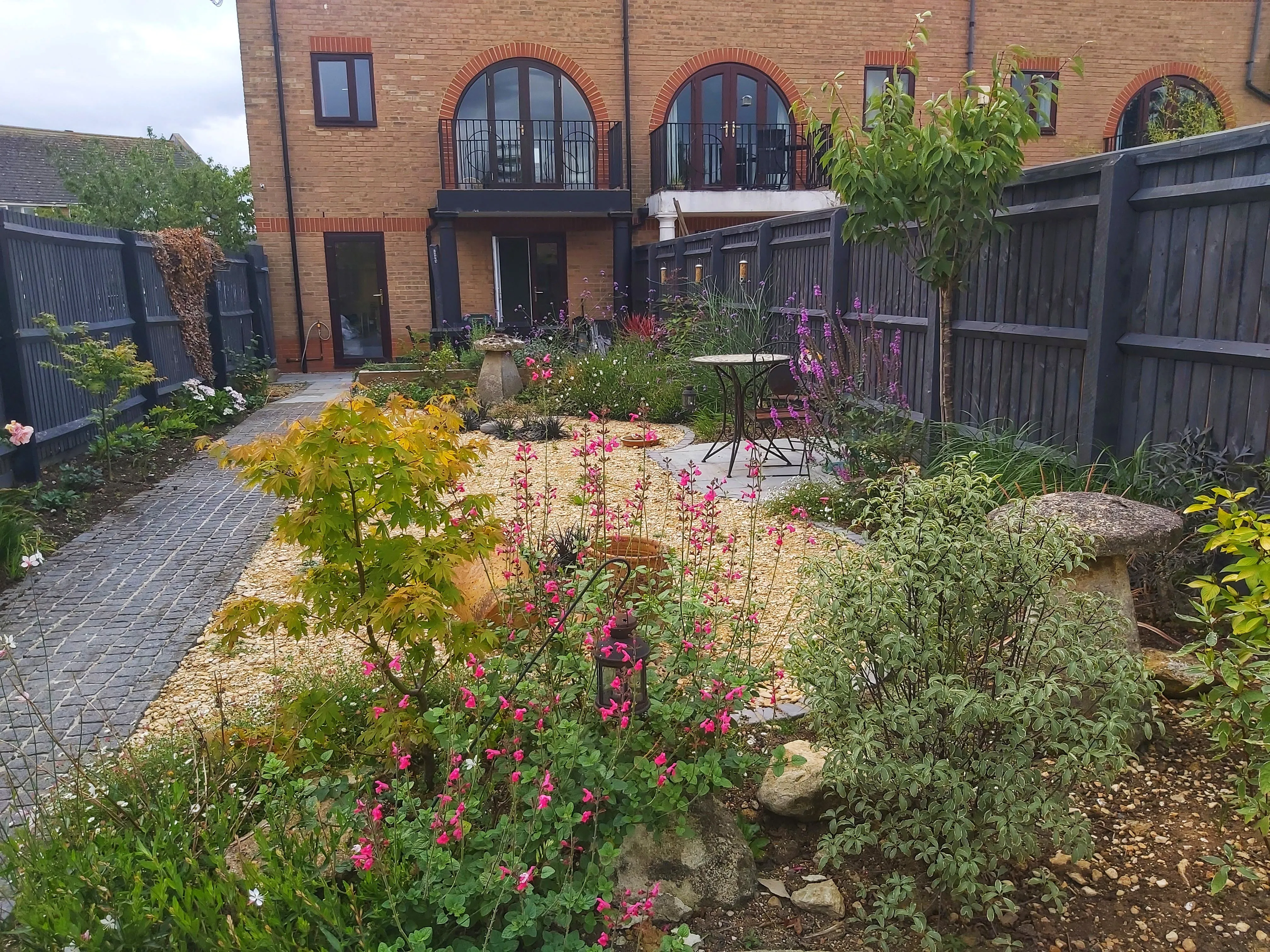 The same view of the house. The grass has been replaced with a bistro set on gravel, surrounded by bright flowerbeds