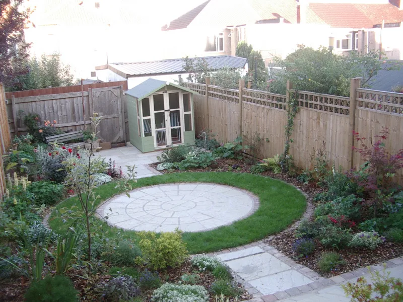 A large circle of slabs surrounded by packed flower beds with a small path leading down to a summer house